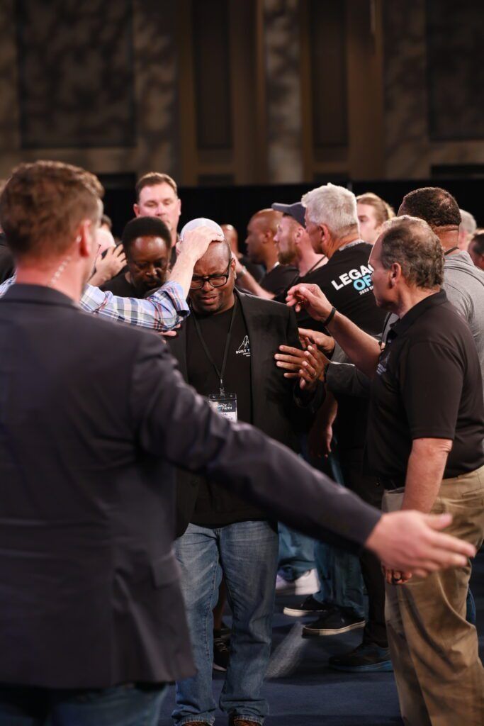 Crowd of people gathered indoors, some talking and gesturing, with one man in the foreground
