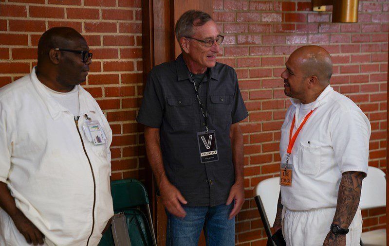 Three people chatting indoors against a brick wall, two in white shirts and one in a dark shirt with a lanyard.