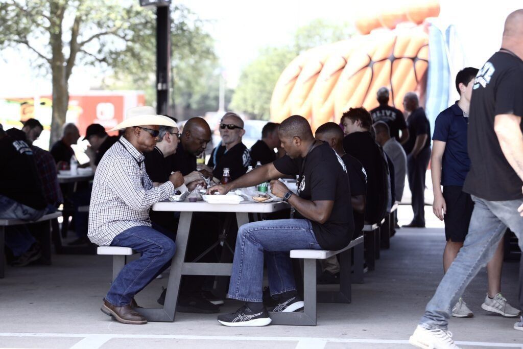 People seated at picnic tables under a shaded outdoor area, eating and talking.