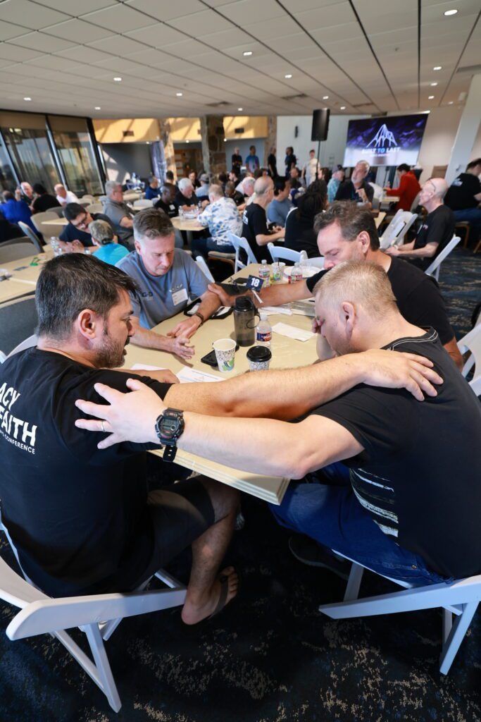 People seated at long tables in a busy indoor event hall, talking and socializing.