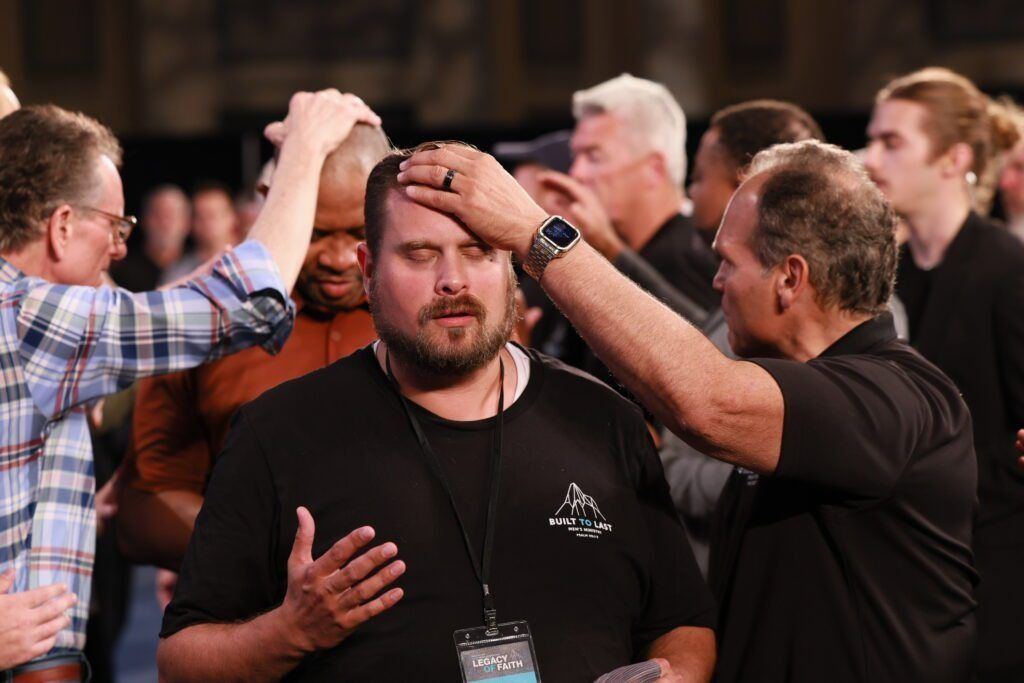 Two men touch a third man’s forehead in a crowded indoor gathering