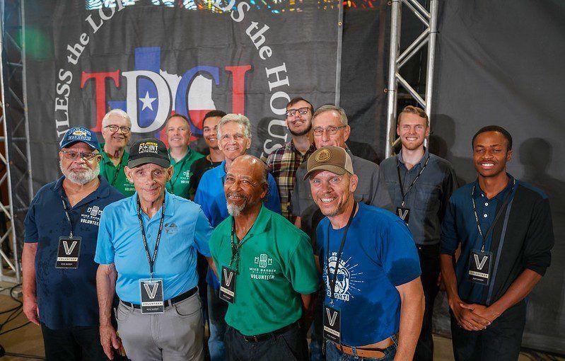 Group of smiling people standing on stage in front of a TDC banner, wearing lanyards and hats.