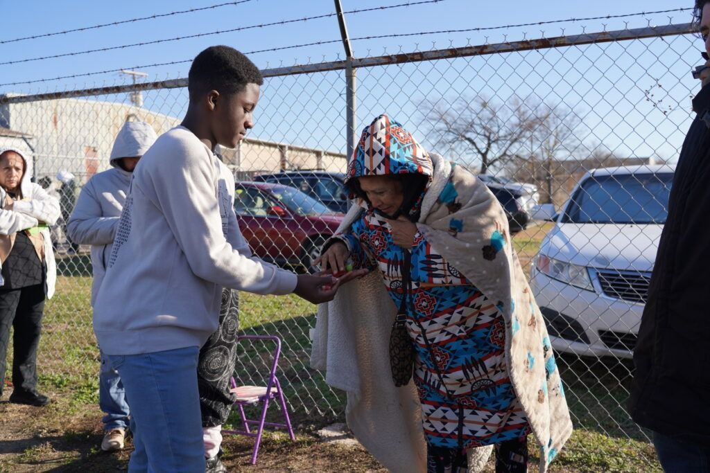 Person hands food to a bundled-up person outdoors near parked cars and a fence.