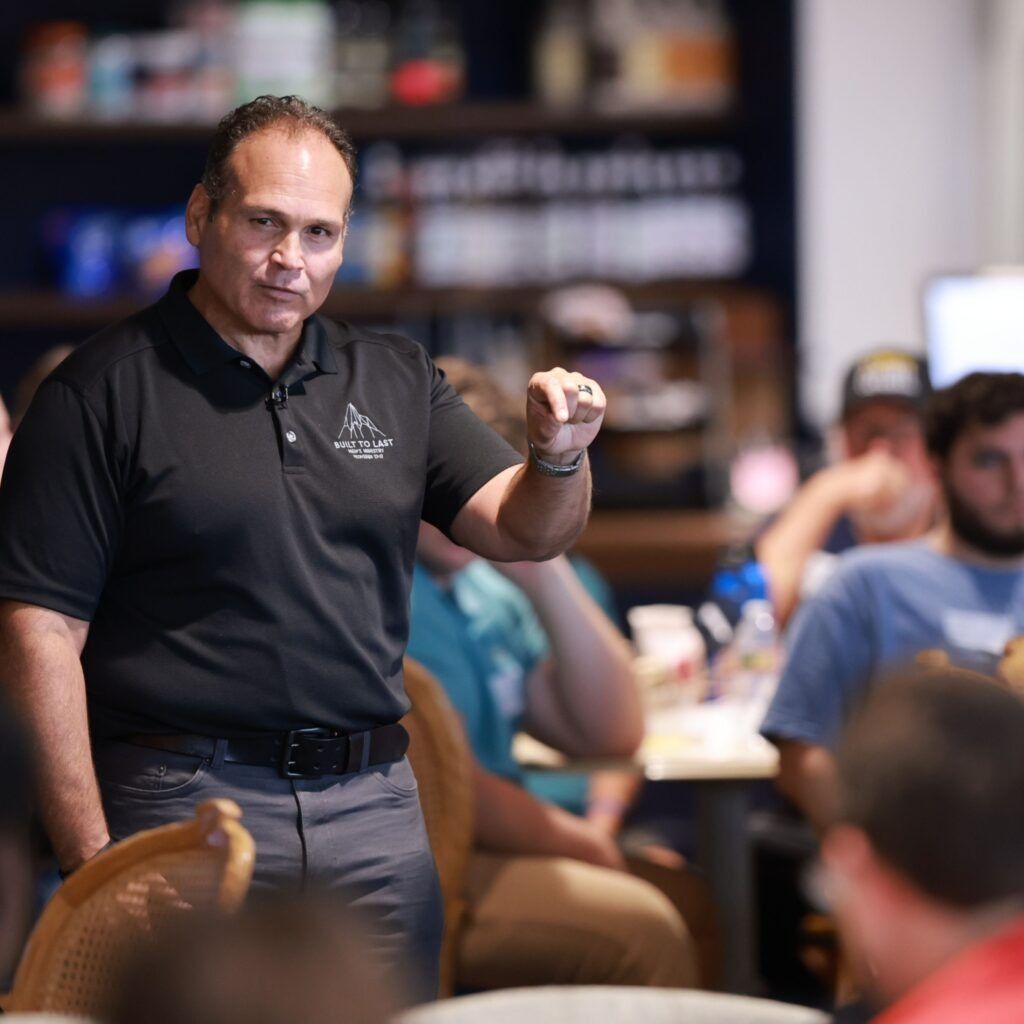 Man speaking to a seated audience in a conference room, gesturing with a raised hand.