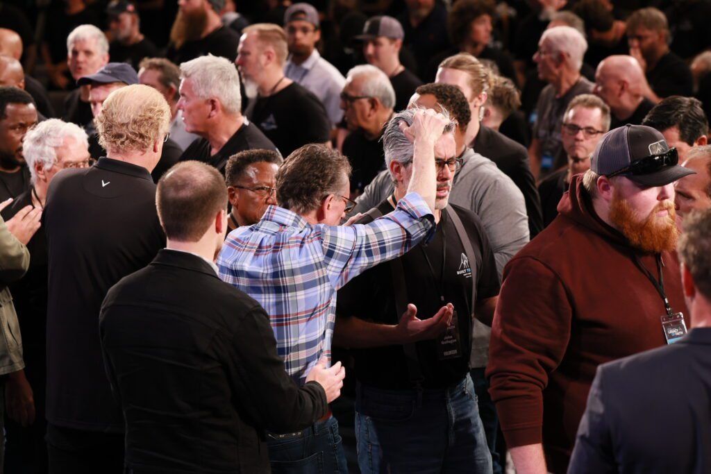 Crowd of people indoors, one in a blue plaid shirt raising a hand toward the stage