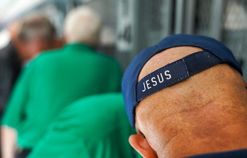 Back of a person wearing a blue cap with “JESUS” on it, seated in a blurred crowd