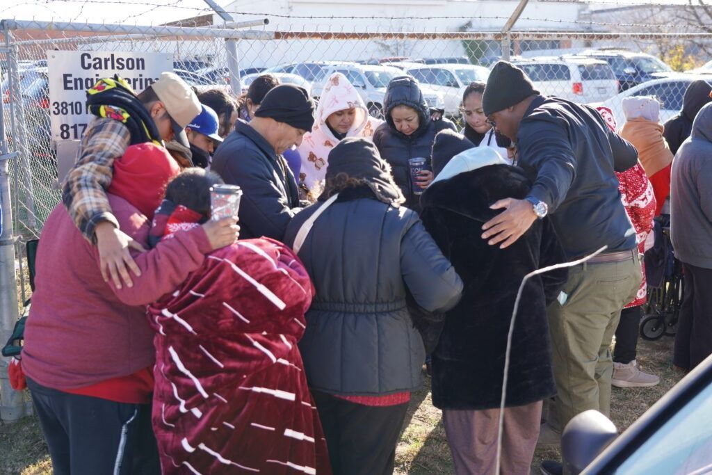 Crowd of people in winter coats and hats gathered outdoors near parked cars, huddled in a group