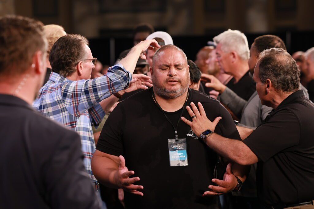 People gathered in a crowded room, one man in black looks overwhelmed as others touch his shoulders.