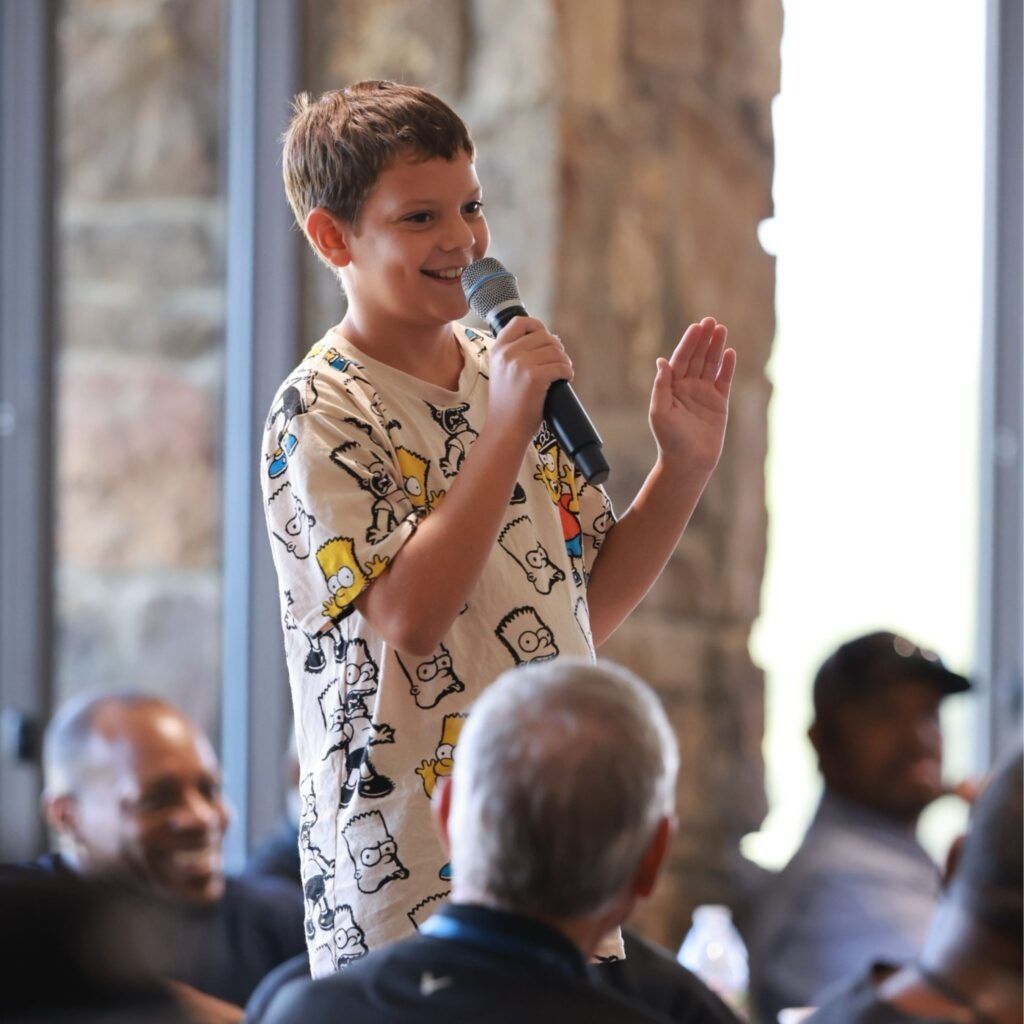 Boy speaking into a microphone at an indoor event, smiling and gesturing to the audience.