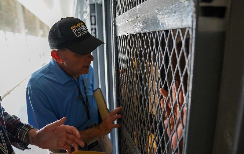 Man in a blue shirt and black cap speaking through a chain-link gate to a person inside a cell