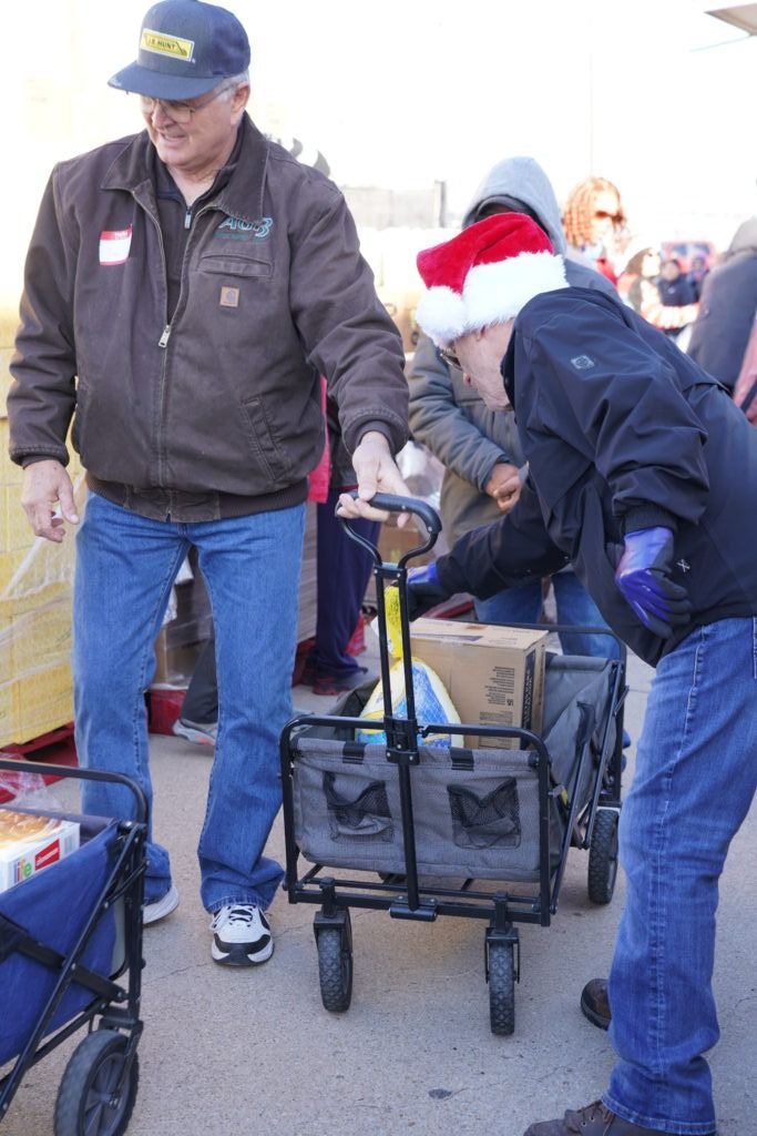 Two people loading a black wagon with supplies in an outdoor parking lot, one wearing a Santa hat