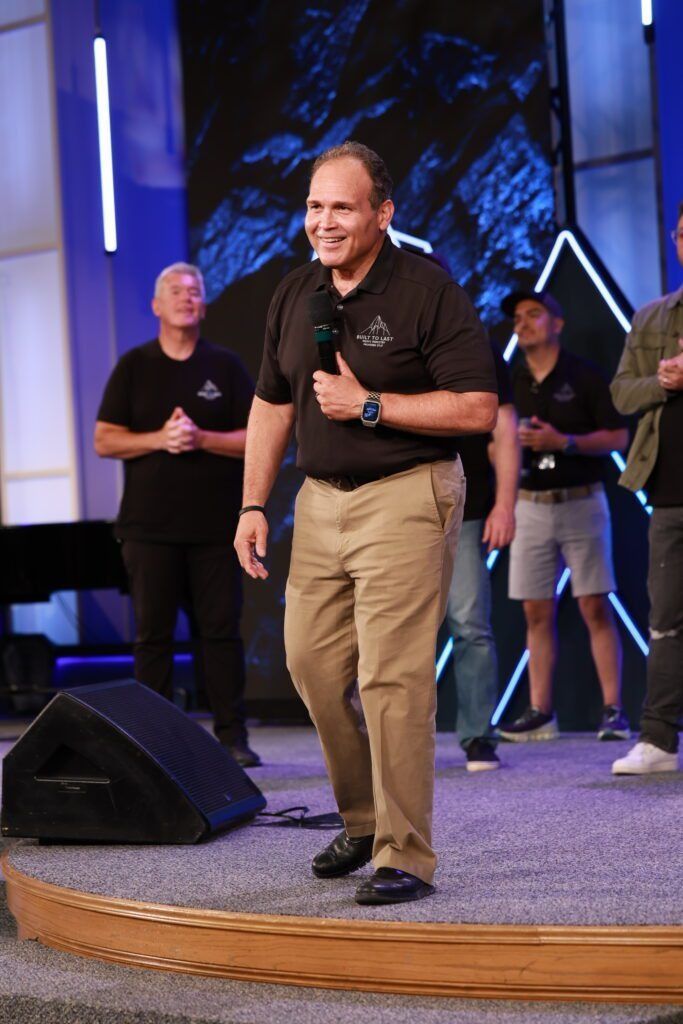 Man speaking on a stage with a microphone, wearing a black shirt and khaki pants, with people standing behind him.