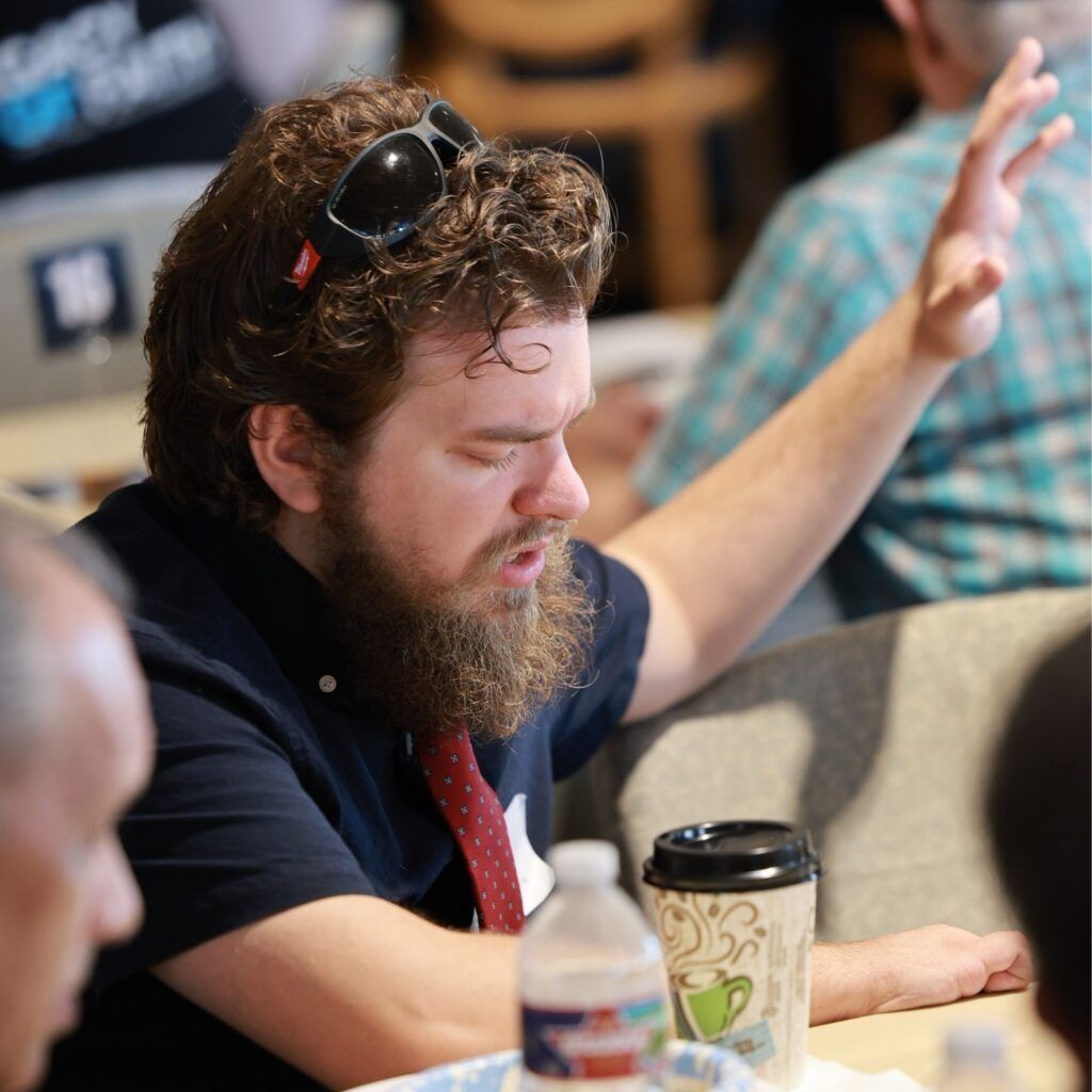 Bearded man in a dark shirt gestures while speaking at a table with drinks, in a casual indoor setting