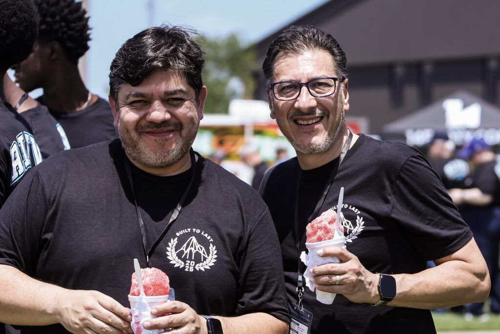 Two smiling men in black shirts hold shaved ice cups outdoors at a sunny event.