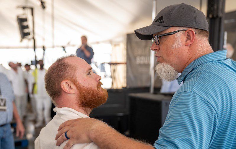 Two men speaking face to face at an outdoor event, one with a hand on the other's shoulder.