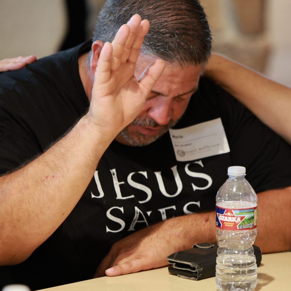 Man with eyes closed raises hand in prayer beside water bottle and phone, wearing “JESUS SAVES” shirt