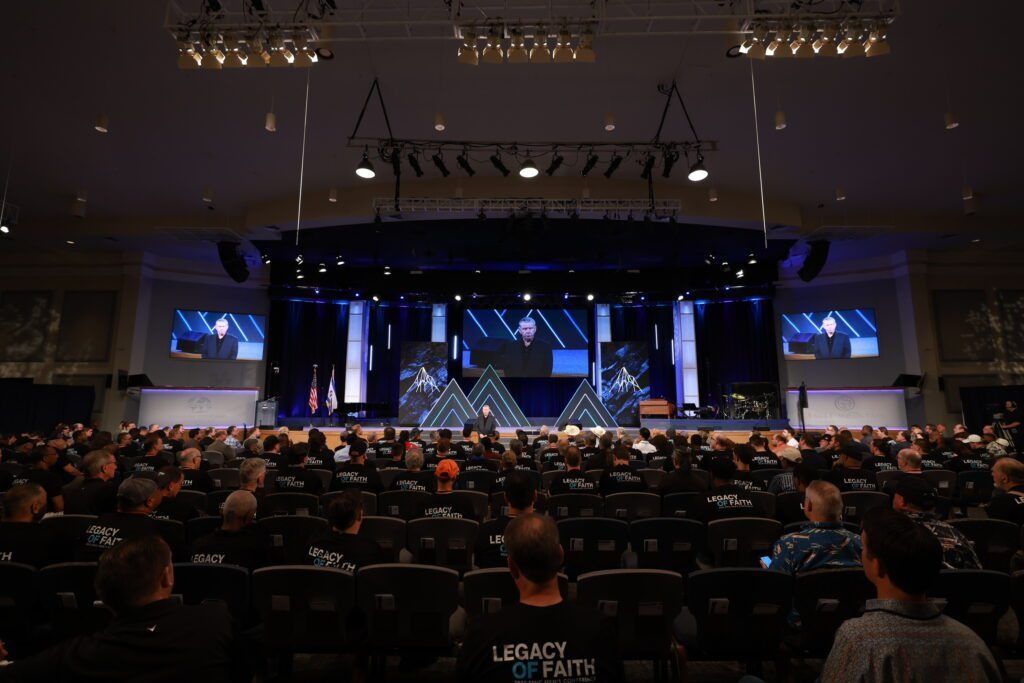 Large conference auditorium with a crowd facing a lit stage and multiple screens