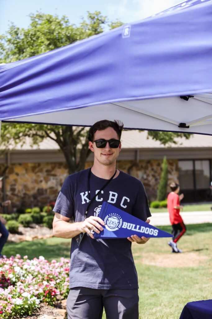 Person under a blue canopy holding a blue banner outdoors, wearing sunglasses and a navy T-shirt.