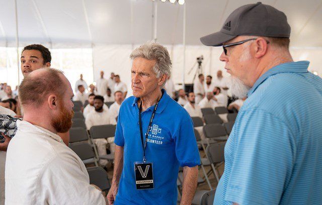 Three men talking in a bright conference room, one wearing a blue polo and event badge.