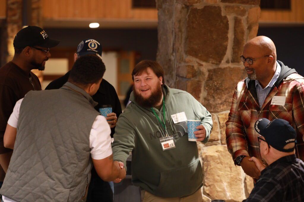 Five people chatting indoors near a stone column, one holding a cup and lanyard badge.