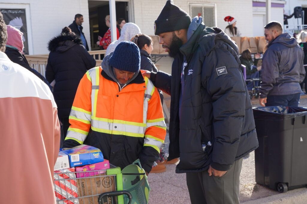 A volunteer in an orange safety jacket helps a shopper with groceries at an outdoor market