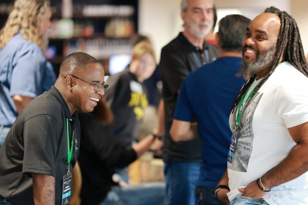 Two smiling men chatting in a busy indoor event space, one in a gray shirt and one in a white shirt.