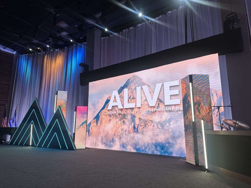 Large stage with “ALIVE” screen, blue lighting, and geometric LED décor in a convention hall