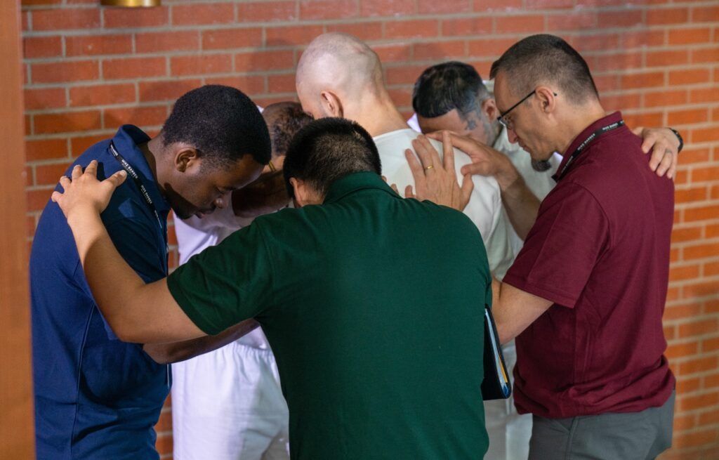 Five people stand in a huddle with heads bowed, arms around each other in prayer or support.