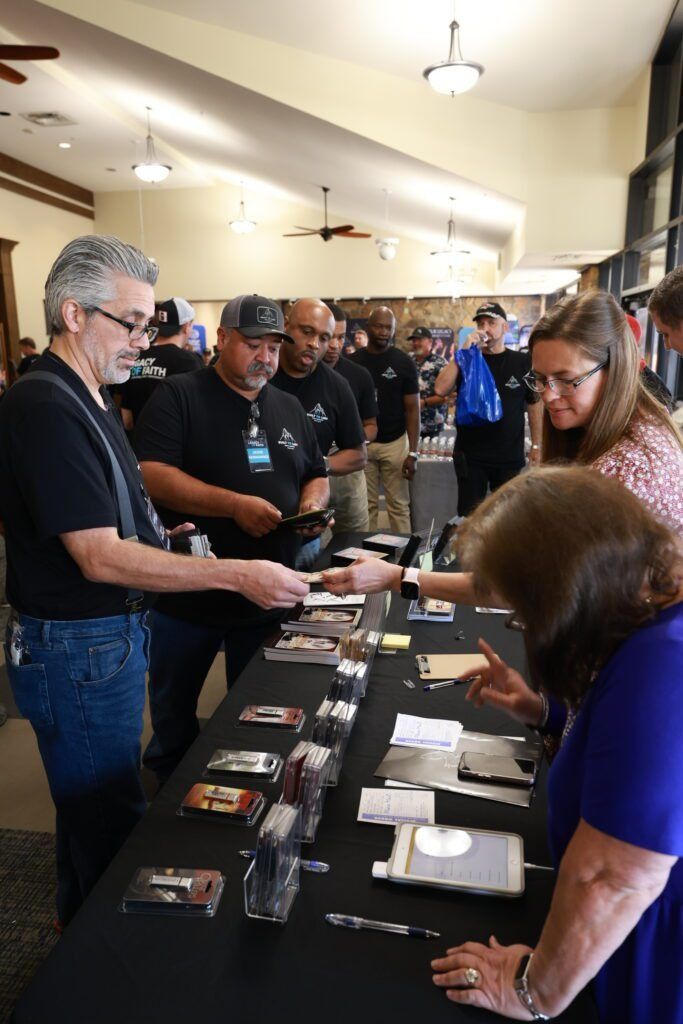People in black shirts serving items at a long table in a crowded indoor event hall.