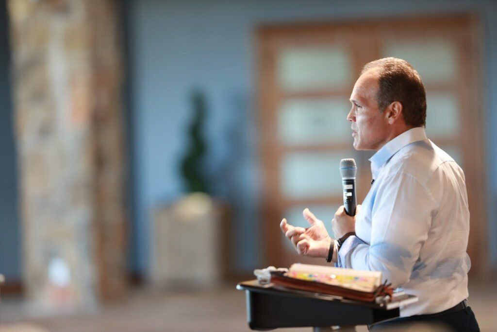 Man speaking into a microphone at a podium in a conference room, with papers in front of him