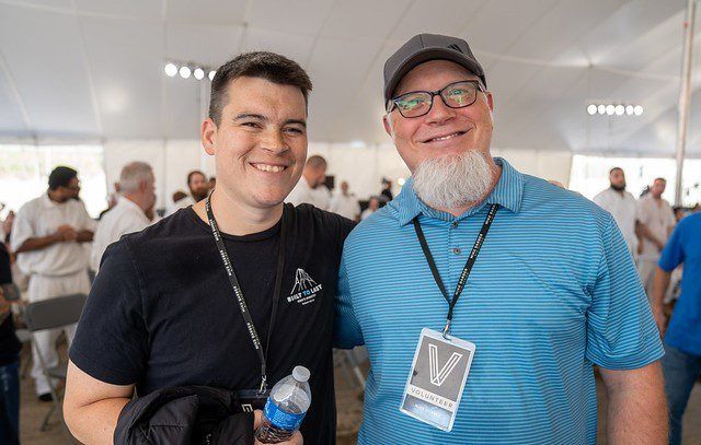 Two smiling people at an indoor event, one holding a water bottle and both wearing lanyards.