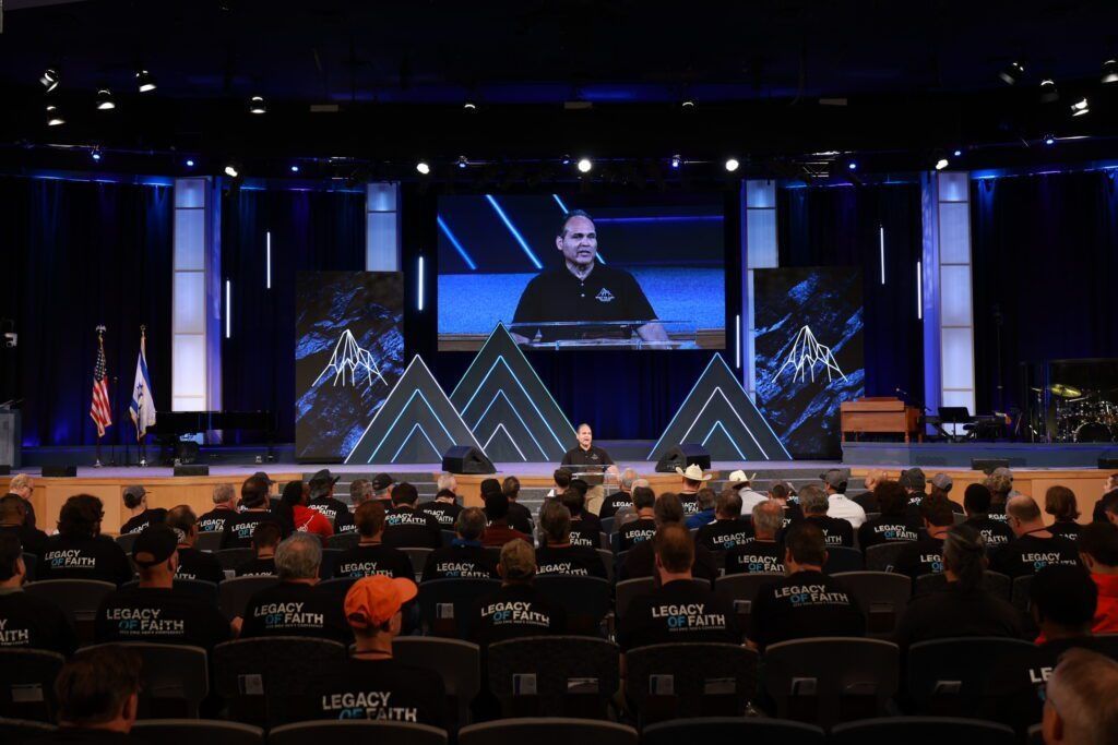 Auditorium full of attendees facing a stage with blue lighting and a speaker on a large screen