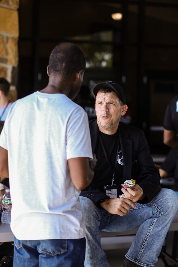 Two people talking outdoors, one seated holding a can, the other standing in a white T-shirt.