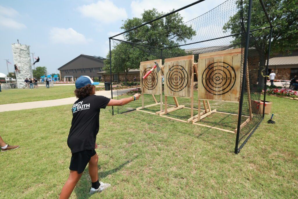 Person throws a tomahawk at wooden targets on a fenced grassy field.