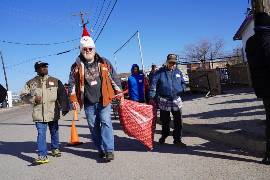 Four people walk down a street carrying a large red sack, one wearing a Santa hat.