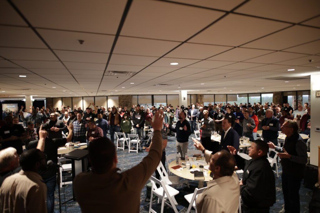 Large crowd gathered in a banquet hall, many standing with raised hands under a low ceiling.