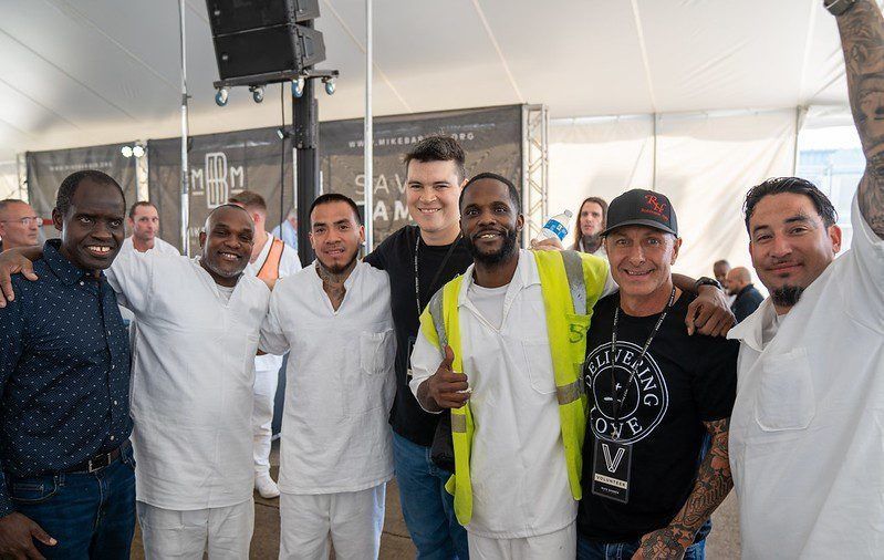 Group of smiling people posing together indoors, one holding a bottle and another wearing a neon safety vest.
