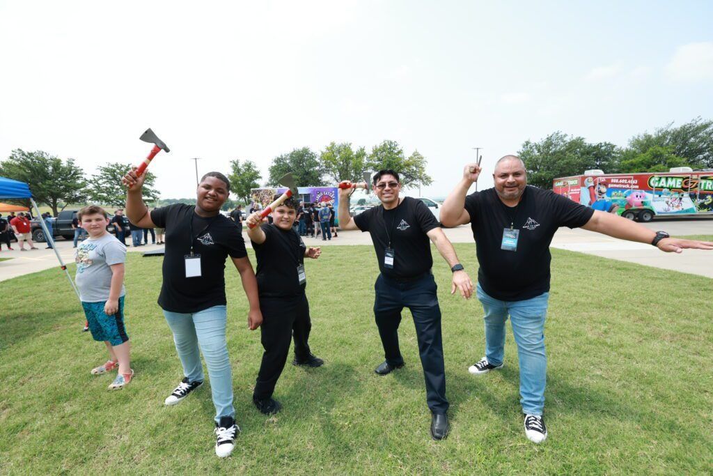 Five people cheering and posing with hammers on a grassy field at an outdoor event