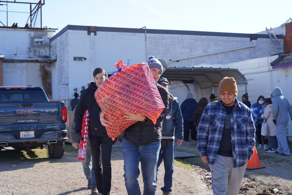 People carrying blankets and bags walk through a sunlit outdoor lot beside a building.