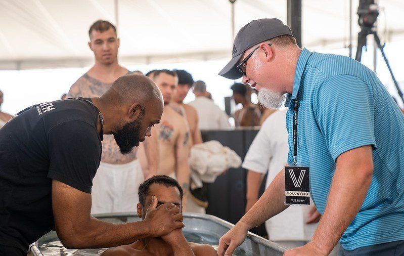Two men help a seated person in a tent, with a crowd and camera equipment in the background.