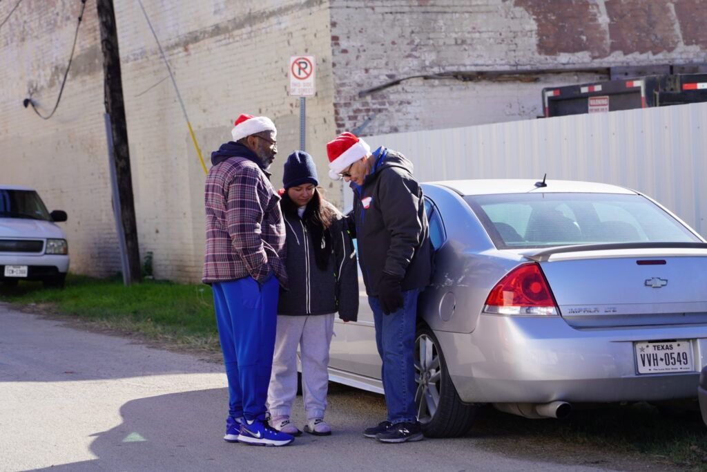 Three people in Santa hats stand beside a silver car in a parking lot.