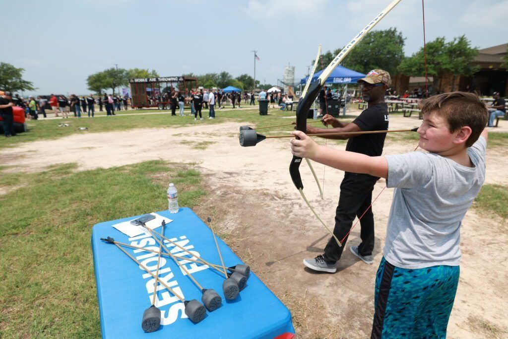 Boy aiming a black bow at a target in an outdoor archery event, with spectators in the background