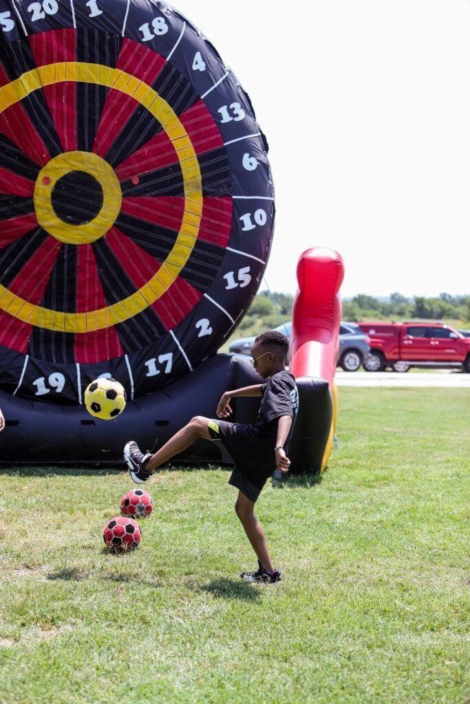 Child kicking a soccer ball toward a giant bullseye target on a grassy field near a red inflatable dummy