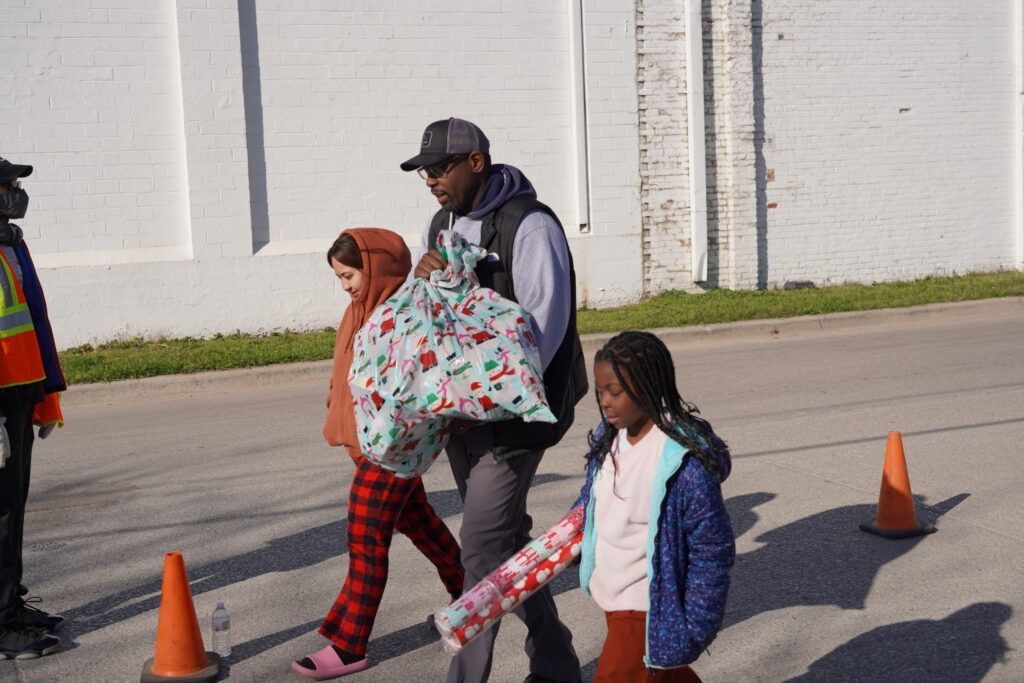 Children walking outdoors near orange cones, one carrying a patterned blanket and another holding a bag.