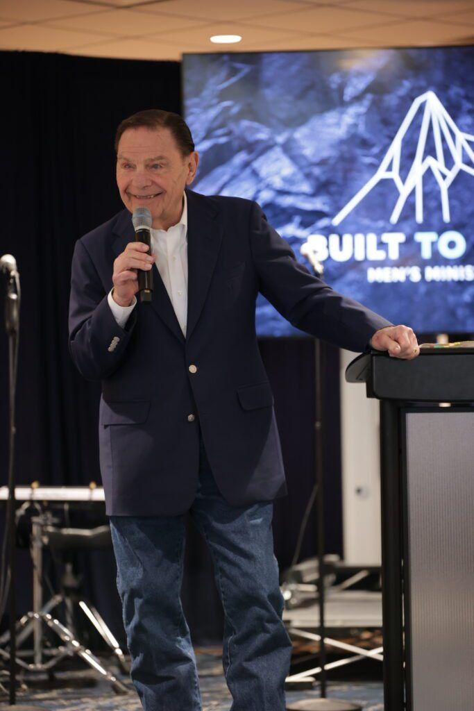 Man speaking into a microphone beside a podium in a banquet hall, with a “Built to...” banner behind him