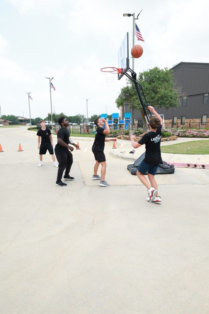 People playing outdoor basketball on a paved court, one jumping to shoot near orange cones and flags.