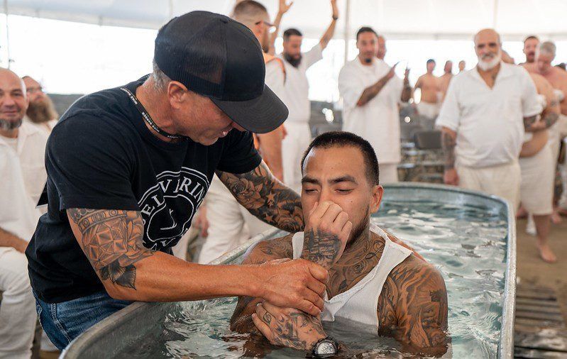 Man baptizing a man in a pool, with onlookers standing in the background.