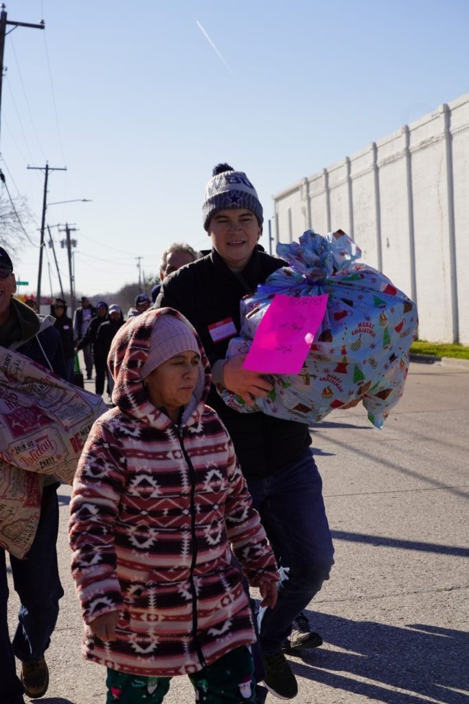 Two children on a sunny street, one carrying a large gift-wrapped bundle with a pink tag