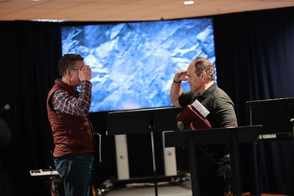 Two people facing each other on a stage, one holding a book, in front of a large screen.