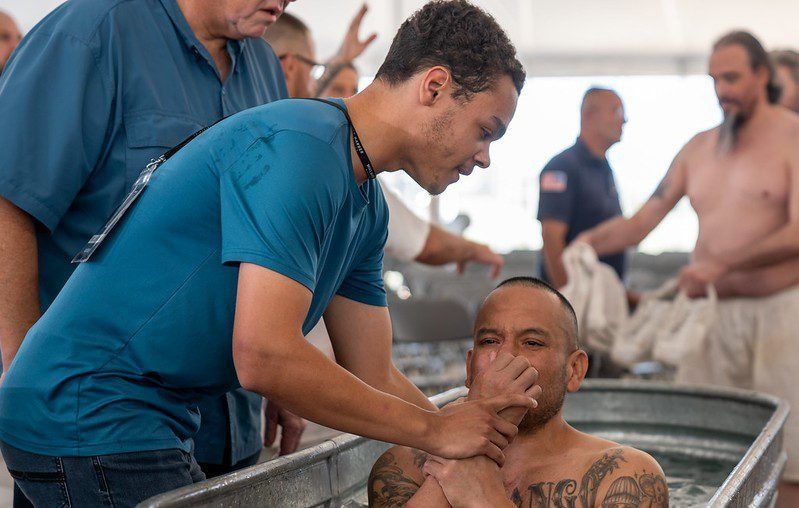 A man in a blue shirt assists another man being baptized in a metal tub during a church ceremony.